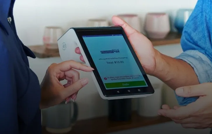 A payment terminal with a PromptPay payment screen, held out to a customer who taps the screen